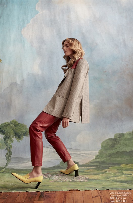 Fashion editorial shot of a fair-skinned adult female in red leather pants and a blazer, posing against a painted landscape backdrop.