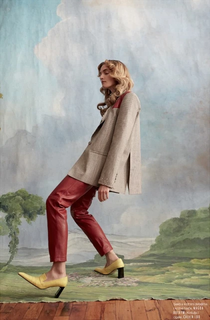 Fashion editorial shot of a fair-skinned adult female in red leather pants and a blazer, posing against a painted landscape backdrop.