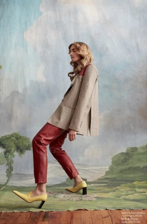 Fashion editorial shot of a fair-skinned adult female in red leather pants and a blazer, posing against a painted landscape backdrop.