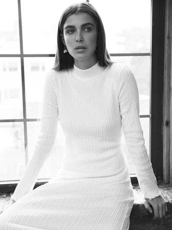 Portrait of a woman wearing a ribbed long-sleeve dress seated against a large grid window in a monochrome setting
