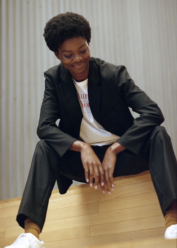 Stylish adult female in a blazer sitting indoors on a wooden surface smiling, fashion shoot, advertising campaign. Natural light.
