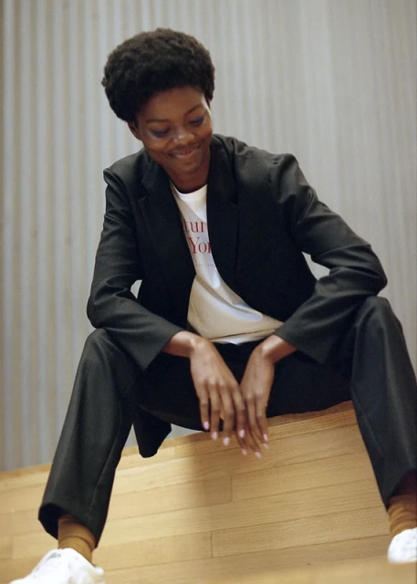 Stylish adult female in a blazer sitting indoors on a wooden surface smiling, fashion shoot, advertising campaign. Natural light.