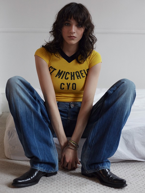 Indoor portrait of a young adult female with long brown hair, wearing a yellow V-neck t-shirt, blue jeans, and black leather shoes.