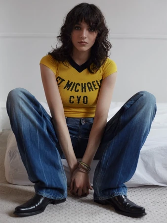 Indoor portrait of a young adult female with long brown hair, wearing a yellow V-neck t-shirt, blue jeans, and black leather shoes.