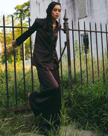 Woman with long braided hair wearing a dark satin pantsuit posing against a metal fence outdoors in Los Angeles.