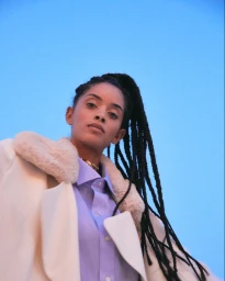 Portrait of a woman with long braided hair wearing a white faux fur collar coat and light blue shirt against a clear sky background
