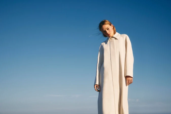 A woman in a long cream white coat standing against a clear blue sky background