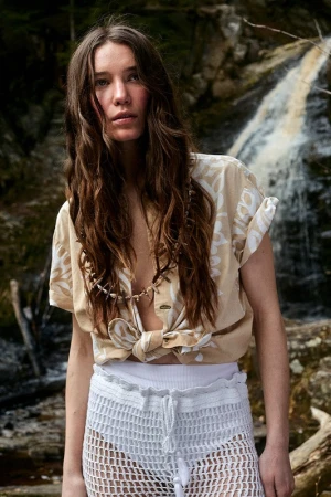 Woman with long brown hair wearing a beige printed shirt and white crochet shorts standing before a waterfall.
