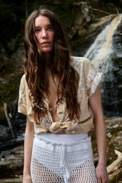 Woman with long brown hair wearing a beige printed shirt and white crochet shorts standing before a waterfall.