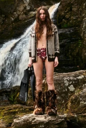 A woman wearing a patterned cardigan and furry boots holds swim fins standing before a rushing waterfall.