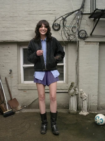 Smiling woman in black quilted jacket, light blue shirt, and purple shorts standing outdoors by a stone wall