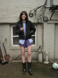 Smiling woman in black quilted jacket, light blue shirt, and purple shorts standing outdoors by a stone wall