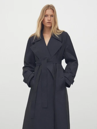 Model with long blonde hair wearing a black trench coat posing against a plain light grey studio background.