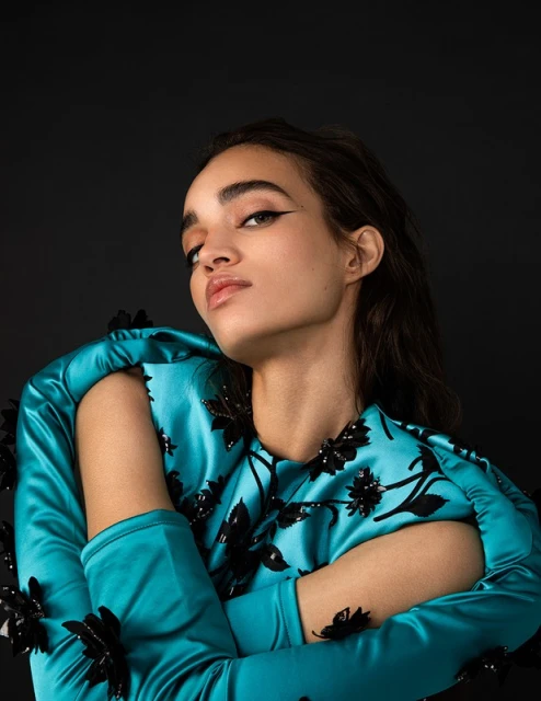 Portrait of a young adult woman with dark hair wearing a teal satin dress with black floral embellishments against a dark background.