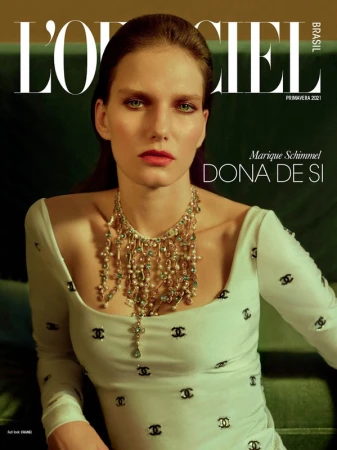 Model wearing a white dress with black logo motifs and an elaborate jewelled necklace against a green background