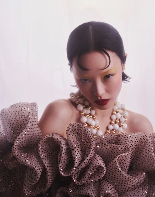 Close-up portrait of a model with dark hair wearing a ruffled dress and a pearl necklace, with yellow eyeshadow, and red lipstick.