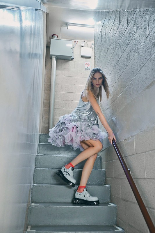 Fashionable young woman in silver dress and chunky shoes poses on gray concrete stairs, holding onto a metal handrail, fashion editorial.