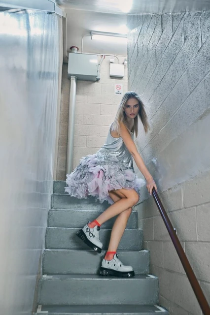 Fashionable young woman in silver dress and chunky shoes poses on gray concrete stairs, holding onto a metal handrail, fashion editorial.
