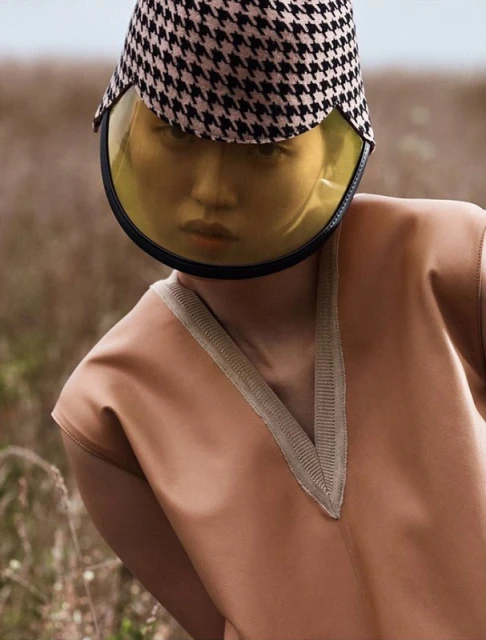 Woman in tan dress and houndstooth sun hat poses in a field. The hat has a clear yellow visor. Fashion editorial photograph.