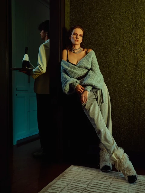 Elegant woman in a sweater leans against a green wall, while a server with a wine bottle stands in the doorway. Studio lighting.