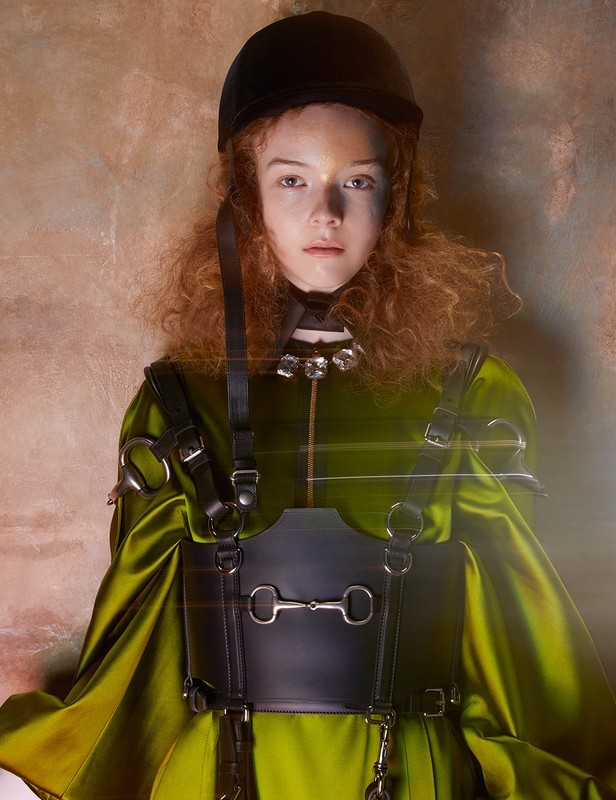 Portrait of a curly red-haired person wearing a black equestrian helmet and green satin dress with decorative black horse bit harness.