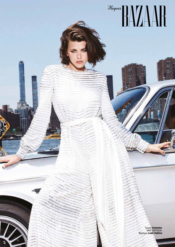 Editorial fashion shot of a woman in a white jumpsuit standing next to a vintage car, with city buildings in the background under blue sky.