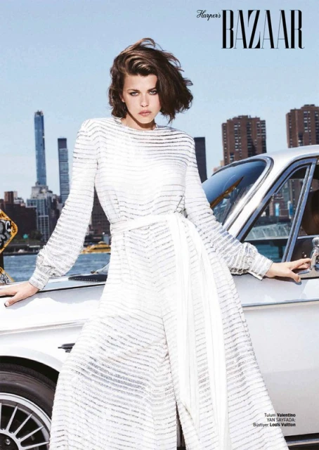 Editorial fashion shot of a woman in a white jumpsuit standing next to a vintage car, with city buildings in the background under blue sky.