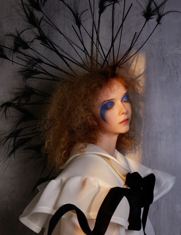 Portrait of a stylish young woman with bright blue makeup, wearing a white ruffled top with a black ribbon and feathered headpiece.