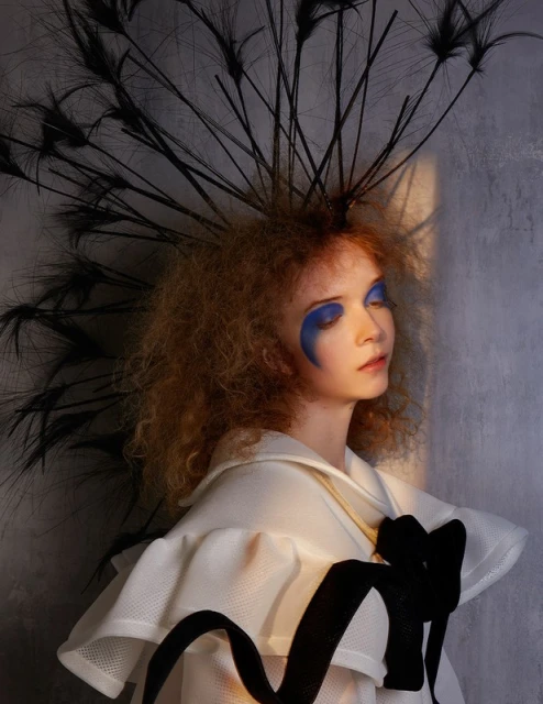 Portrait of a stylish young woman with bright blue makeup, wearing a white ruffled top with a black ribbon and feathered headpiece.