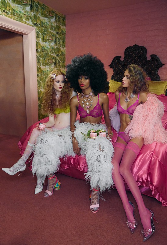 Three women in lingerie sitting on a pink bed holding desserts, featuring a pink bedroom with a patterned green wallpaper accent wall.