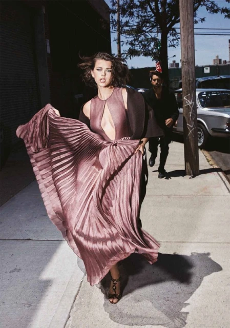 A woman in a mauve gown walks on a sidewalk; a man in a black shirt and party hat trails her on a city street on a sunny day.