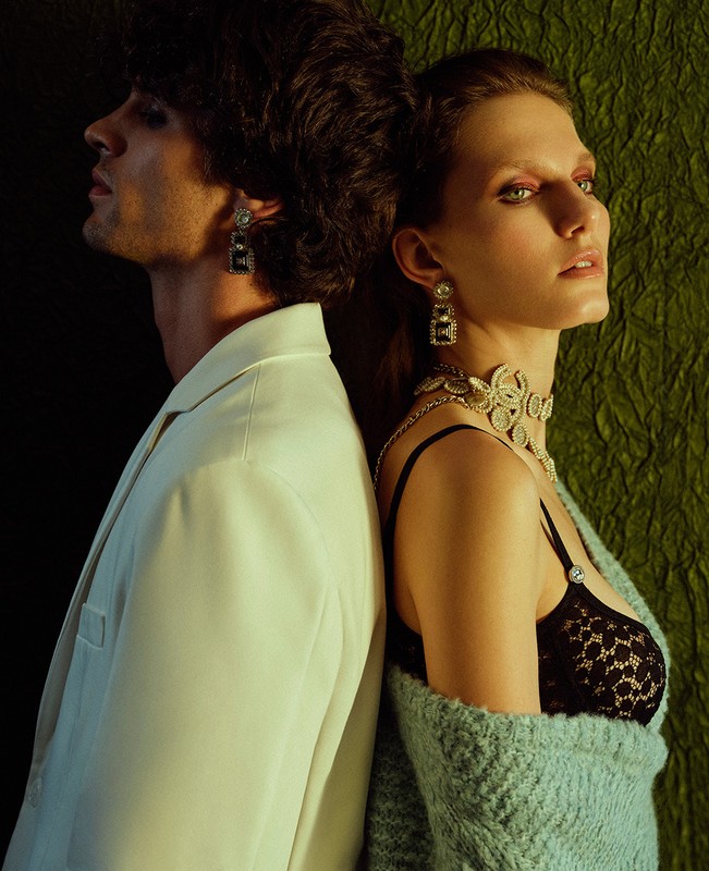 Fashion shot of a man and woman back-to-back in studio wearing elegant jewelry and evening wear against a dark green backdrop.