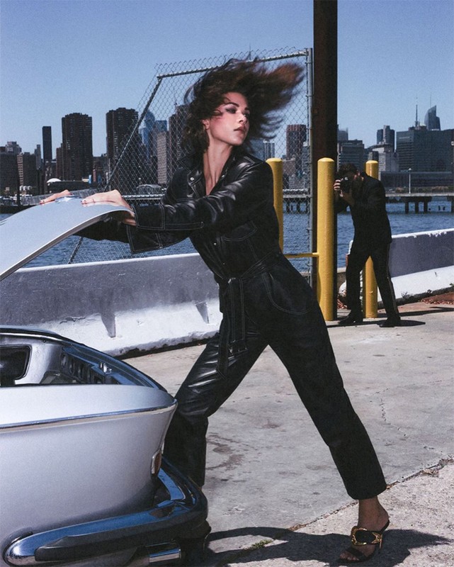 A model in a black leather jumpsuit poses near a silver car on a pier, with a photographer and city skyline in the background.