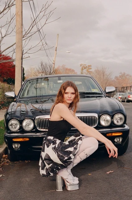 Adult woman crouching in front of a black car on a city street, wearing a black top, patterned skirt, platform shoes, and white stockings.
