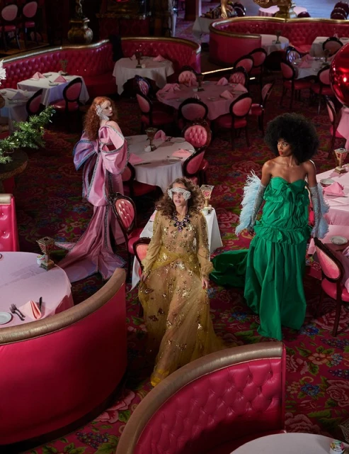 Three women in formal gowns pose inside a retro-style restaurant with pink booths, tables set with pink linens and silver tableware.