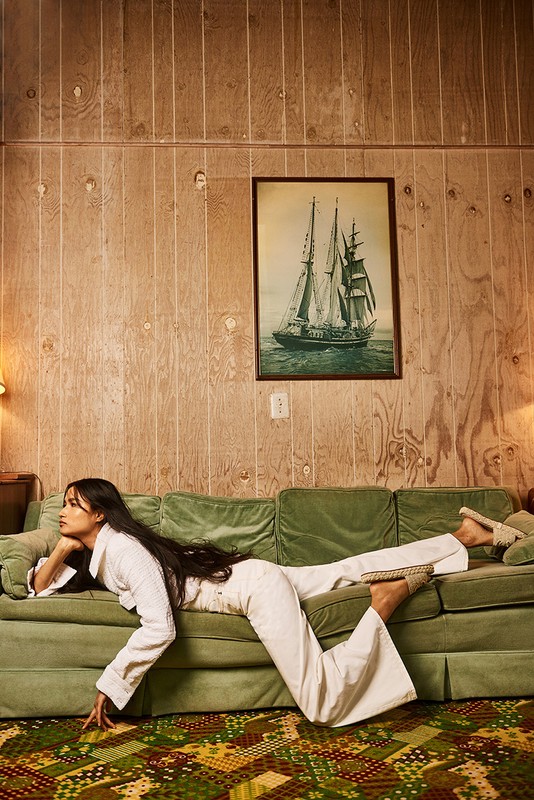 A woman in white loungewear poses on a green velvet couch in an indoor setting with wood-paneled walls and a ship painting.