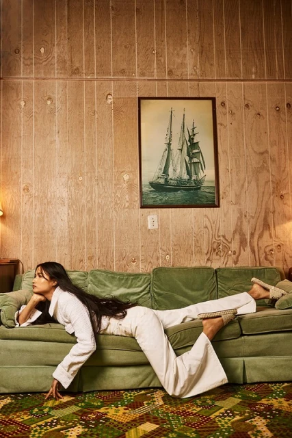 A woman in white loungewear poses on a green velvet couch in an indoor setting with wood-paneled walls and a ship painting.