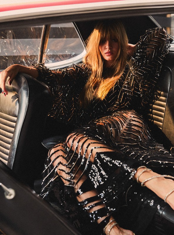 A woman in a sequined dress is sitting in the back of a classic car. Low-angle shot. Natural light and shallow depth of field.