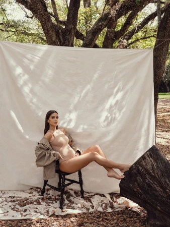A woman in a body suit sits on a black chair in an outdoor setting, with a white backdrop and natural, filtered light from the trees.