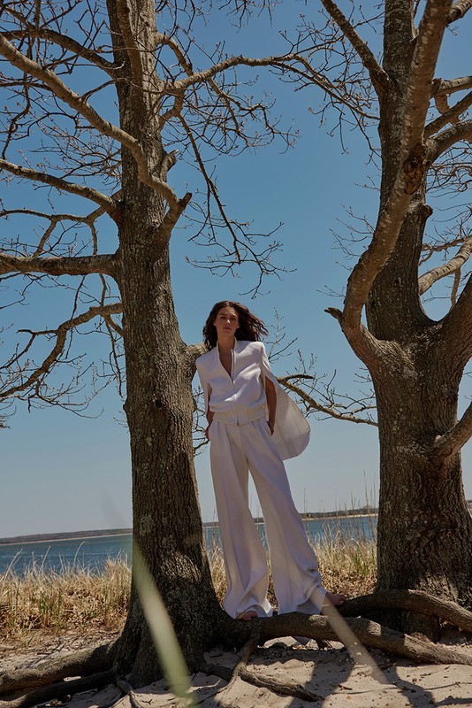 A woman in white attire stands between two trees, gazing slightly off-camera against a backdrop of a light blue sky and ocean water.