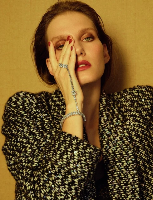 Brunette adult woman poses in black-and-white tweed jacket with silver bracelet and ring against a tan backdrop in a portrait.
