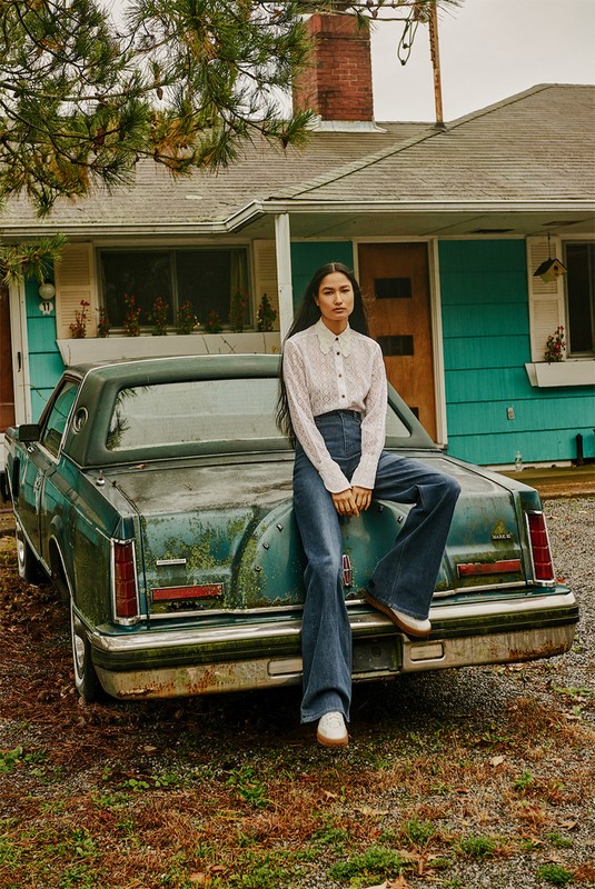 A woman sits on the trunk of a vintage green car parked in front of a turquoise and white house on a cloudy day.