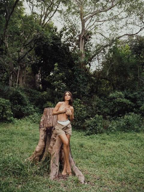 Semi-nude woman poses near a tree stump in a grassy field surrounded by a forest landscape on a sunny day in an outdoor setting.