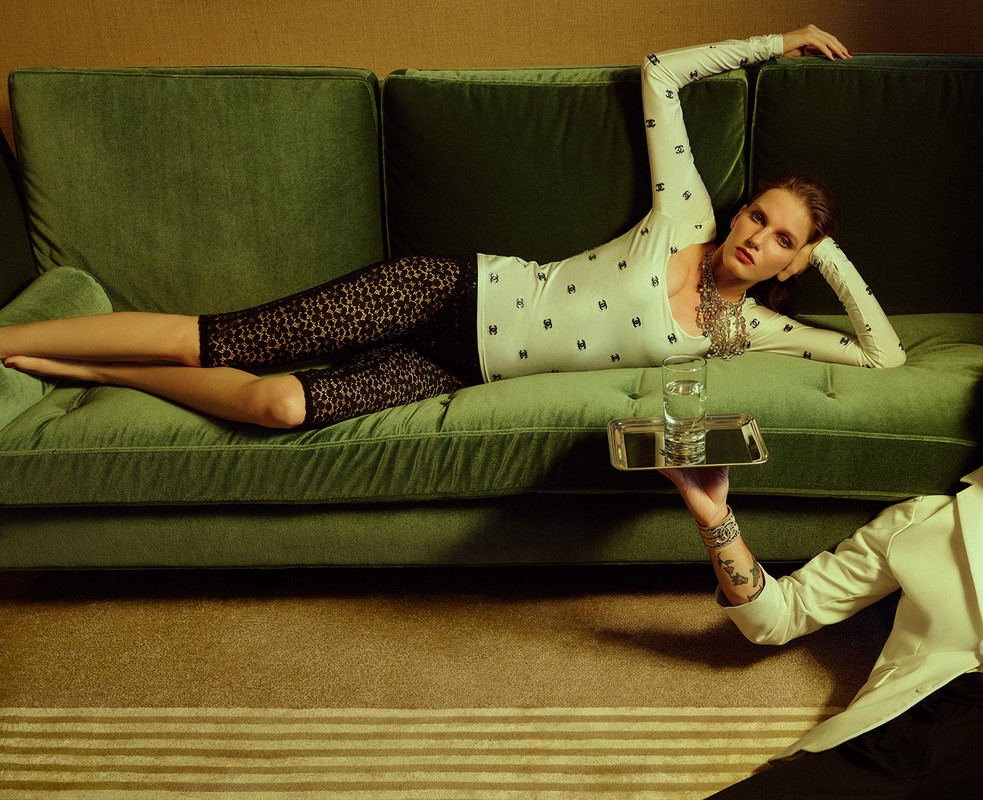 Woman in patterned top and leggings reclines on a green velvet sofa, while a server presents a glass of water on a silver tray.