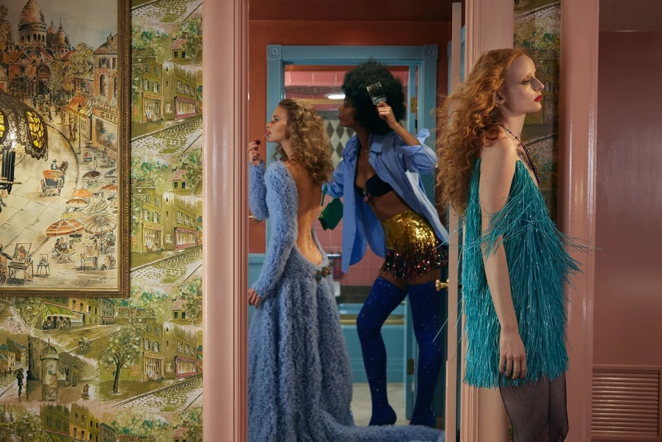 Three women in vintage-inspired dresses are seen through doorways in a vintage-style interior with patterned wallpaper and decor.