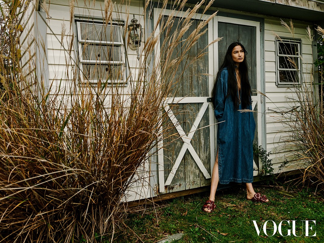 Woman in a denim dress poses in front of a white shed, framed by tall grasses. Vogue logo visible at the bottom.