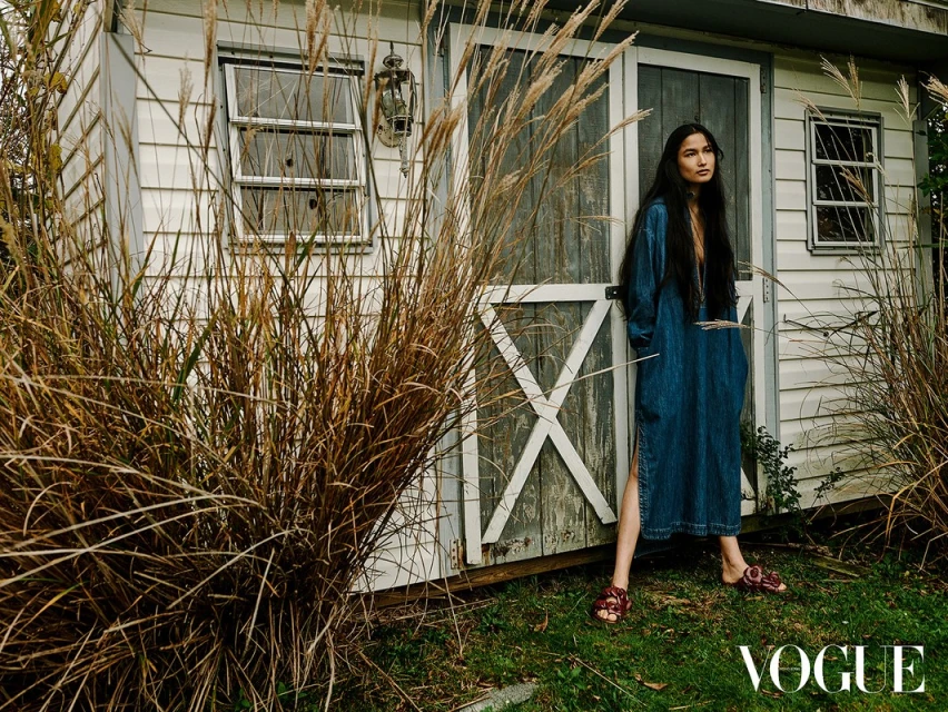 Woman in a denim dress poses in front of a white shed, framed by tall grasses. Vogue logo visible at the bottom.