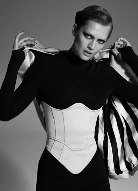 A female fashion model poses in a black long-sleeve dress, cinched with a white corset and striped cape, against a grey backdrop.