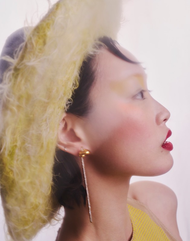 Profile shot of a stylish adult female with bright makeup, a textured hat, and a long earring with rhinestones against a light background.