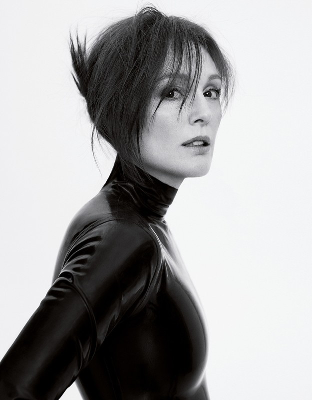 A woman with dark hair in a black latex high-neck top looking over her shoulder against a plain white background